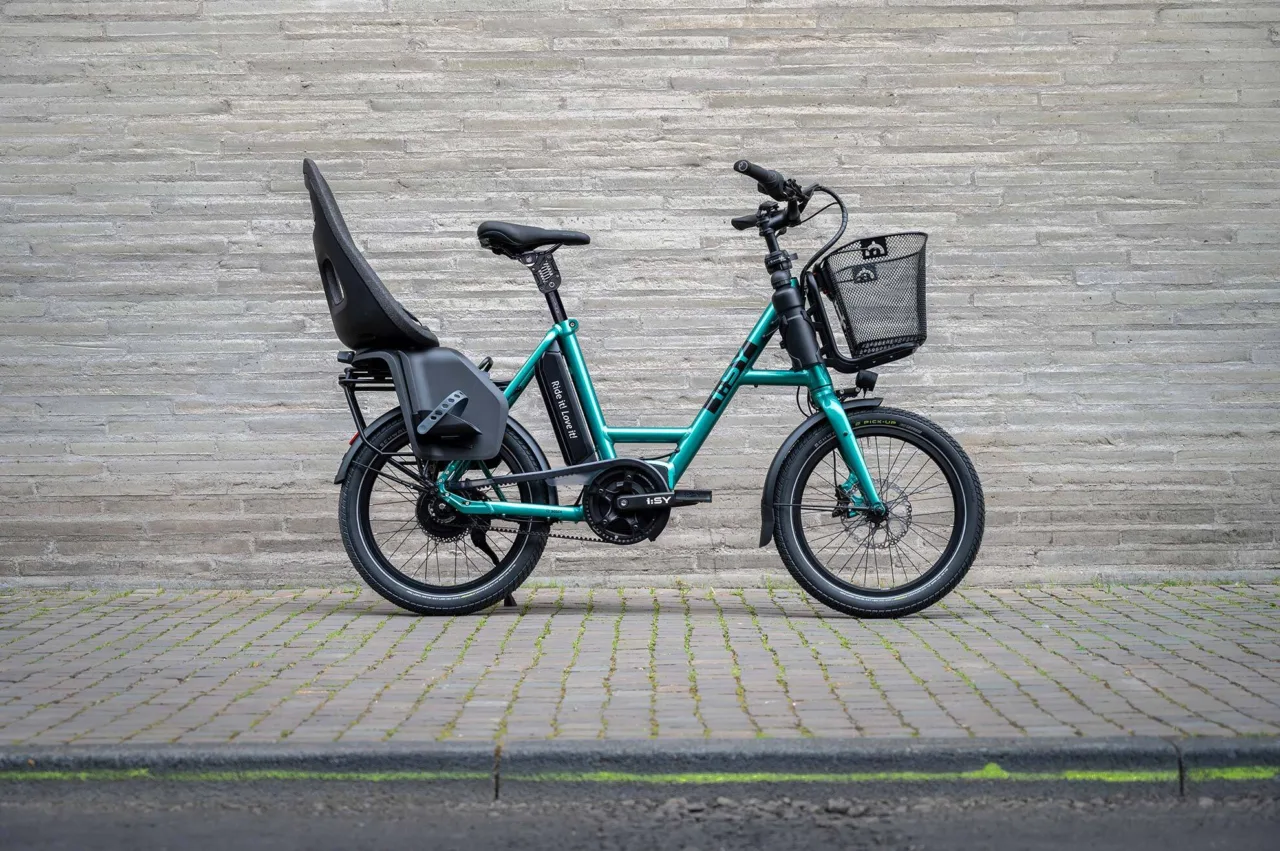Ein kompaktes, türkises i:SY E-Bike mit Kindersitz steht auf einem Bürgersteig vor der Wand eines Gebäudes.